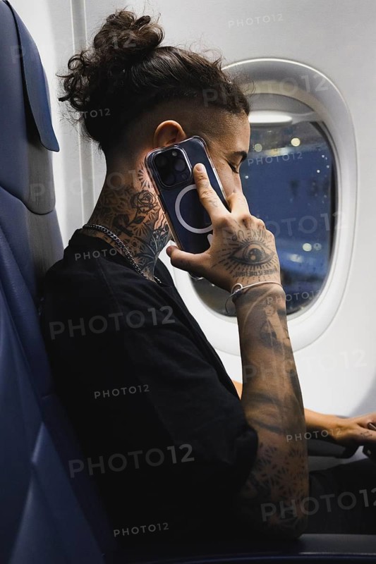 A young woman with detailed tattoos on their arms and neck uses a smartphone while seated on an airplane The scene captures a modern lifestyle with tech and travel themes