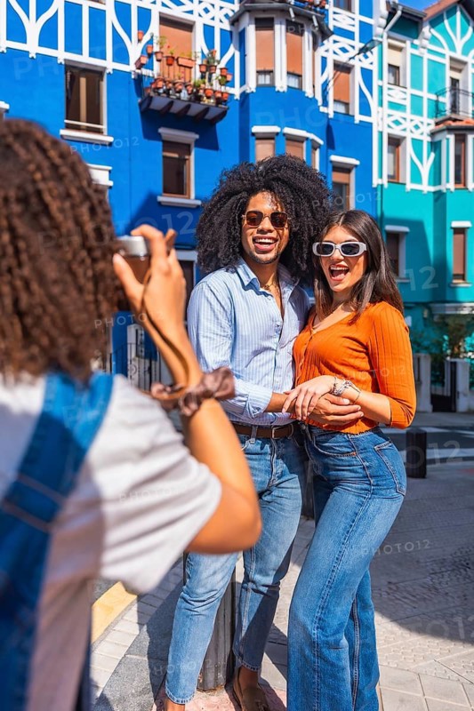 Young photographer taking pictures of a happy tourist couple posing in a colorful street during a sunny day