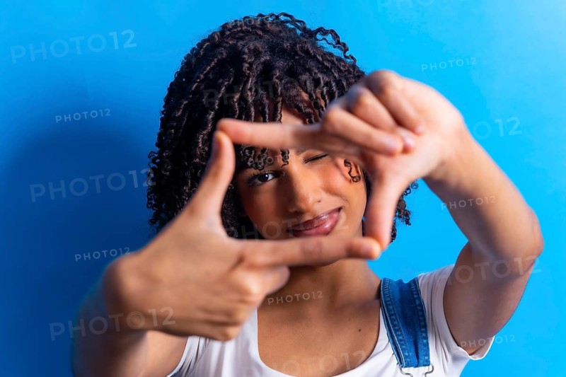 Young woman with curly hair making a frame with her hands around her face, winking, on a blue background