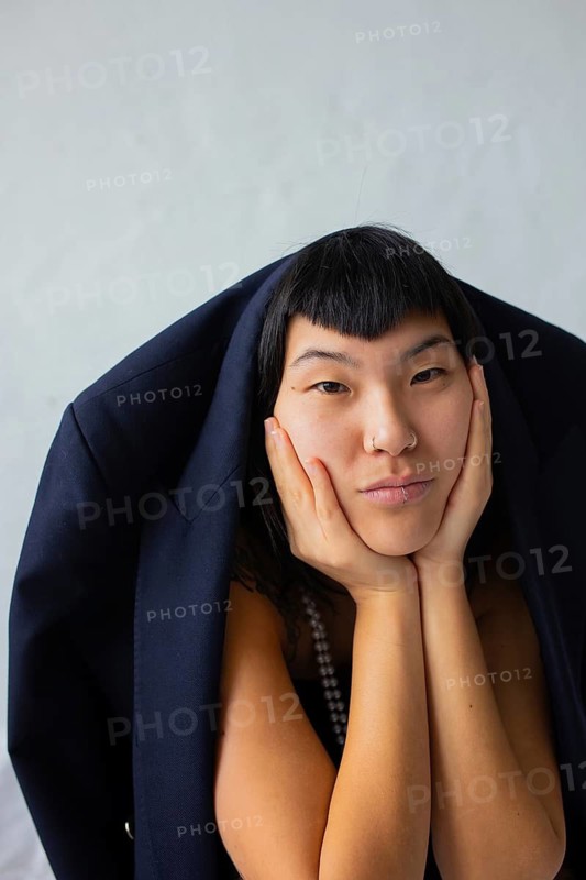 A portrait of a woman with a distinctive hairstyle. Her facial expression is thoughtful, framed by a dark jacket. The minimalistic background complements her features