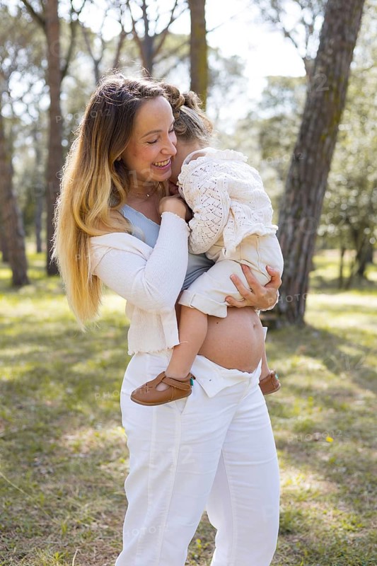 Pregnant woman with her daughter in a serene outdoor setting, sharing a tender moment. The warmth of the sun enhances the peaceful atmosphere in the natural surroundings