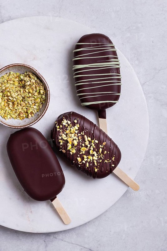 Top view of gourmet chocolate covered ice creams garnished with matcha blend, presented elegantly on a white marble plate, adorned with chopped crushed pistachios, served on a marble surface