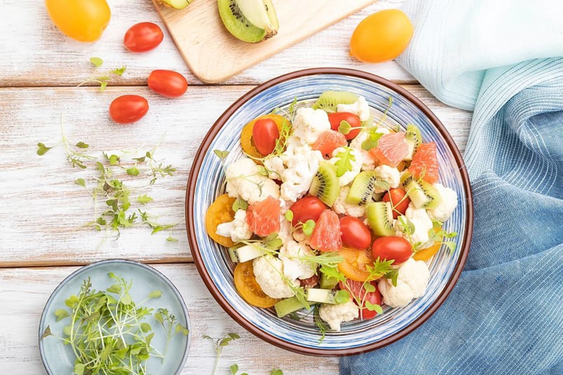 Vegetarian salad of cauliflower cabbage, kiwi, tomatoes, microgreen sprouts on white wooden background and blue linen textile. Top view, flat lay, close up