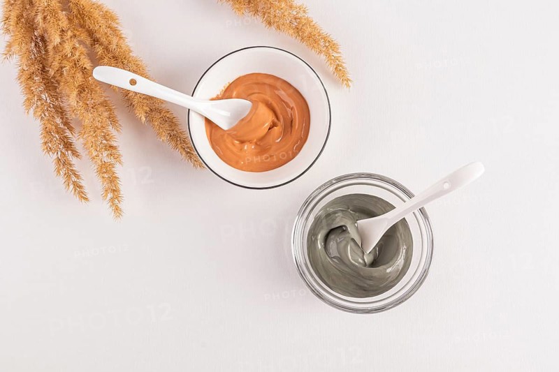 Top view of two bowls of cosmetic clay facial masks, one green and one orange, with white spoons and decorative pampas grass on a neutral background