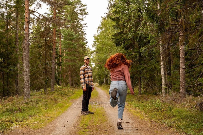 A couple enjoys a leisurely walk along a dirt path in a Swedish forest. The man smiles while the woman joyfully skips, surrounded by lush greenery and tall trees