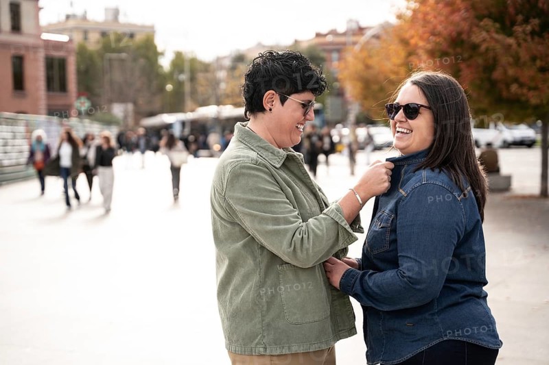 A curvy lesbian couple shares a joyful moment together outdoors, showcasing their unique style and connection. They both wear trendy sunglasses and casual jackets