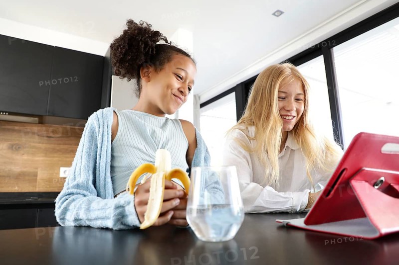 A smiling girl eating a banana and a young woman share a joyful moment in a bright, modern kitchen. They are engaged with a red tablet on the counter, enjoying a relaxed, casual activity