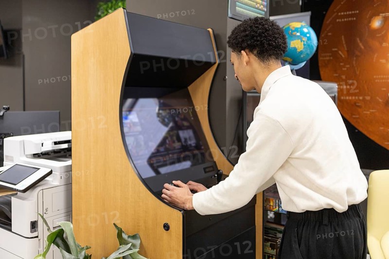 An employee plays on a retro arcade gaming machine during a break in a modern office. Surrounding items include a globe, a printer, and wall posters, creating a relaxed work atmosphere