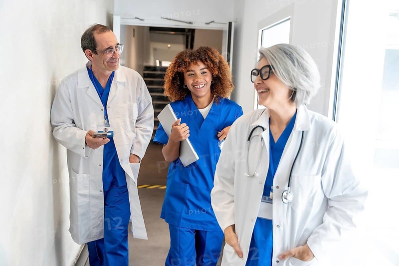 Diverse healthcare team including doctors and a nurse walking together, discussing and smiling in a bright hospital hallway, symbolizing teamwork and positive patient care environment