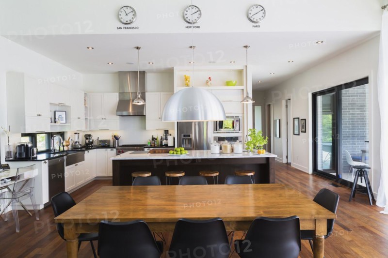 Open-concept dining and kitchen area with antique dining table with black leather and plastic chairs, marble top wooden island with painted black steel and exotic wood bar stools, white cabinets inside a luxurious modern cube style house, Quebec, Canada
