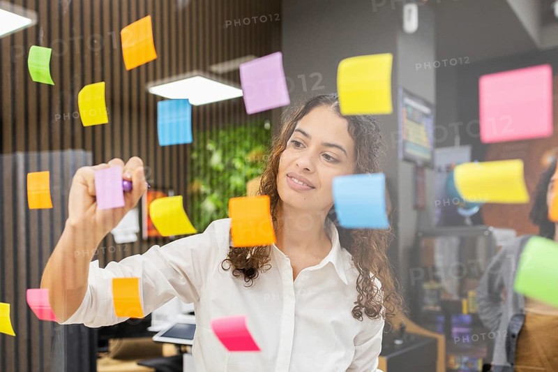 A woman in a modern office uses colorful sticky notes on a glass wall for planning. She's focused and creative, dressed in a white shirt, surrounded by technology and plants