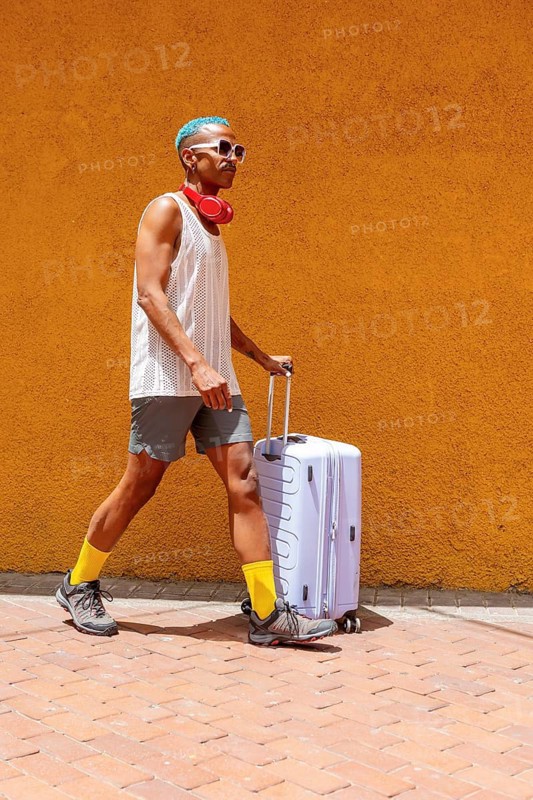LGBTQIA+ person with colorful style walks confidently with a suitcase, preparing for a holiday. Embracing pride and self-expression in a vibrant urban setting