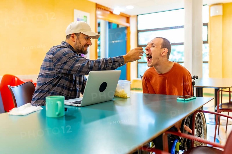 Man feeding a coworker with disability in the public cafeteria of a modern coworking