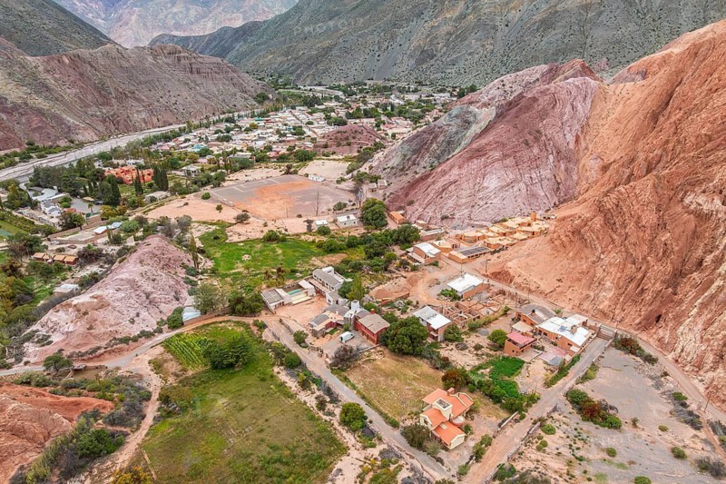 Aerial view capturing the vibrant houses of Purmamarca, Jujuy, nestled against the striking red hills. The picturesque landscape contrasts with lush greenery and rugged terrain