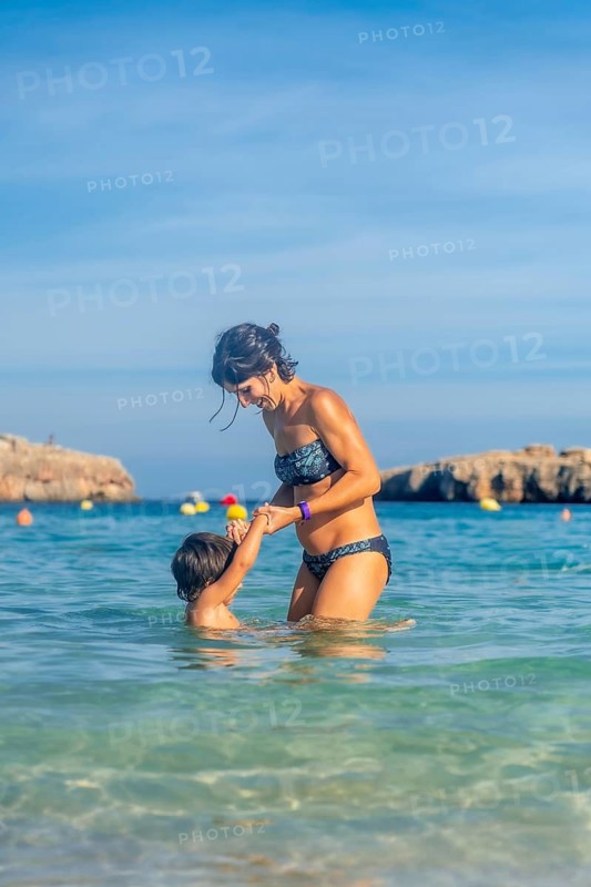 A woman is holding a child in the ocean. The woman is wearing a bikini and the child is wearing a swimsuit. Scene is happy and playful
