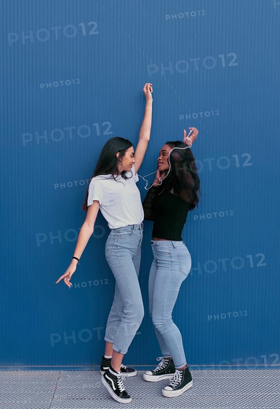 Two young friends share earphones and dance happily against a blue background. They wear casual outfits and sneakers, radiating joy and friendship in their carefree moment