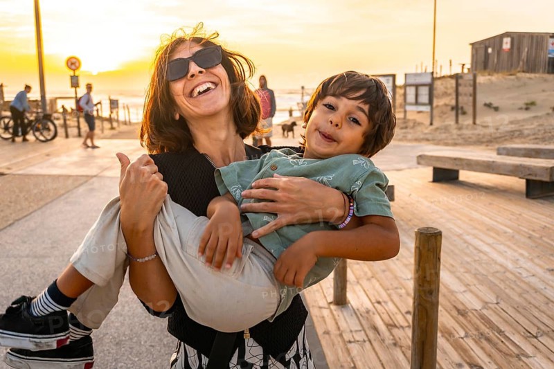 Mother carrying a happy young son, both smiling and making silly faces, enjoying a summer holiday stroll along a wooden boardwalk at sunset on the beach in seignosse, france