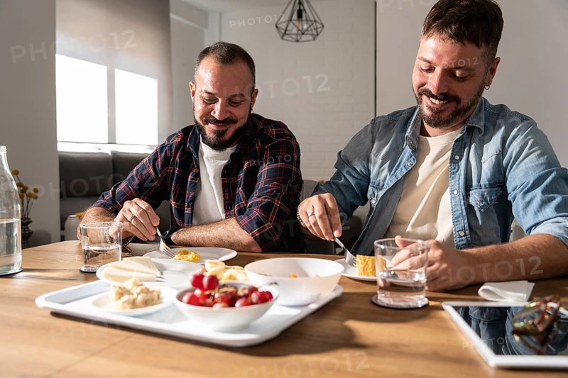 A happy gay couple shares a lighthearted breakfast together in a modern, sunlit kitchen. They are smiling and enjoying each other's company in a warm, relaxed atmosphere