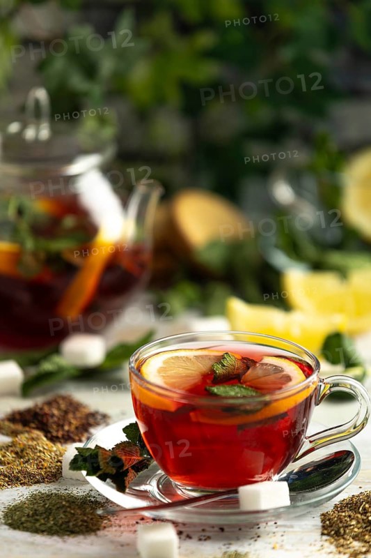 A cup of red tea with lemon and mint, surrounded by teas and sugar cubes, teapot in the background