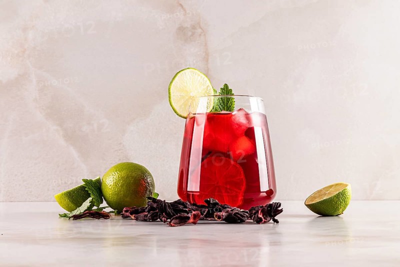A glass of refreshing red hibiscus tea with ice cubes, garnished with a lime slice and mint leaves. Surrounded by fresh and dried ingredients, set on a light marble surface