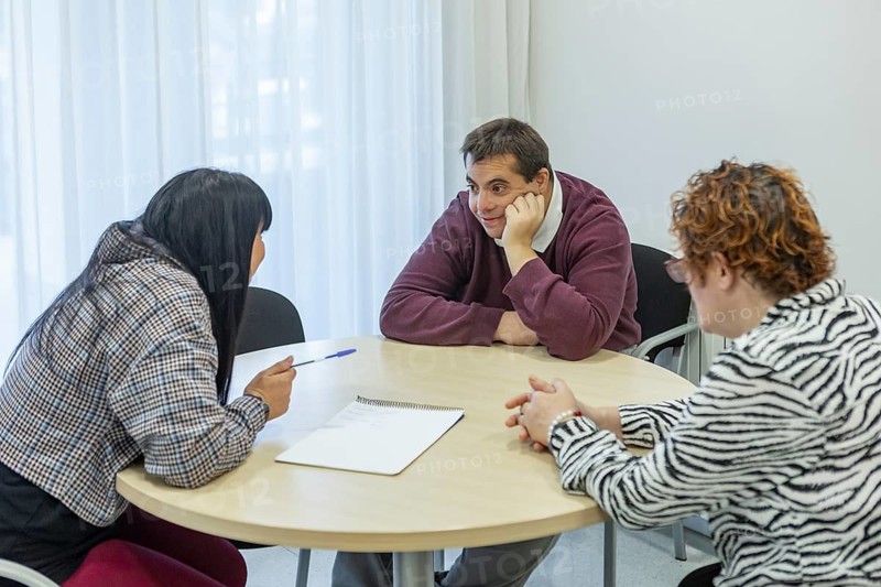 A group of people engage in a discussion in a mental health residence. They are seated around a table, showing interaction and support in a comfortable setting
