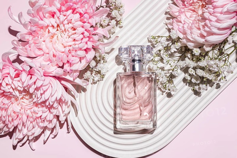 A luxurious perfume bottle rests amidst delicate pink chrysanthemums and white baby breath, creating a sense of elegance and fragrance on a textured white surface