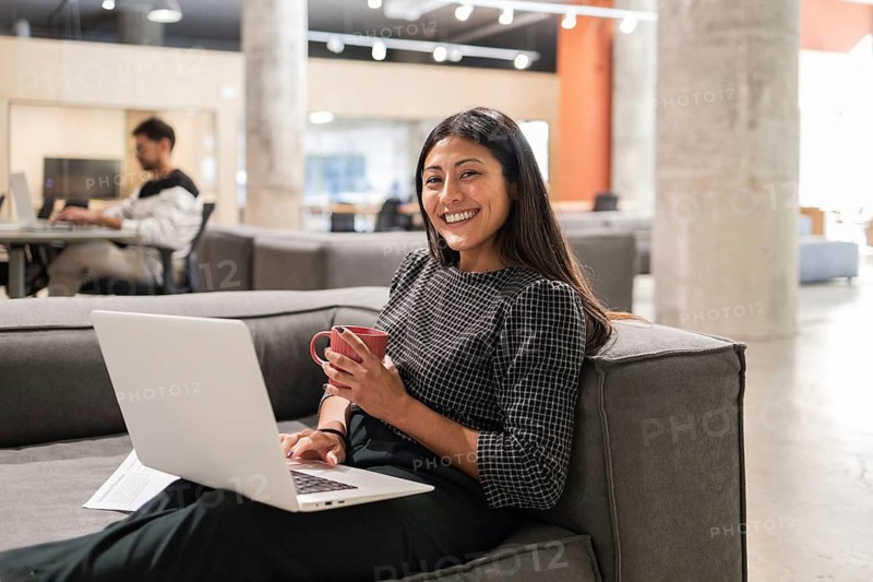 A young professional woman smiles while working on a laptop, holding a coffee mug in a modern office Colleagues work in the background, illustrating a collaborative atmosphere