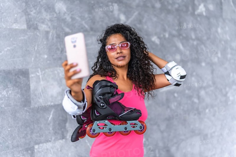 Confident latin woman taking selfie holding inline skates outdoors standing on a urban space