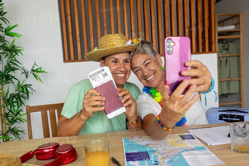 Two joyful lesbians, one in a straw hat, taking a selfie with a passport and tickets, surrounded by maps and travel essentials, planning their summer holiday at home