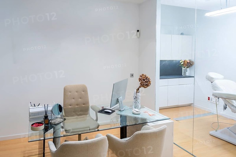 Contemporary aesthetic clinic interior featuring a glass desk workstation with computer and office chairs, alongside a consulting bed in a bright, clean medical environment