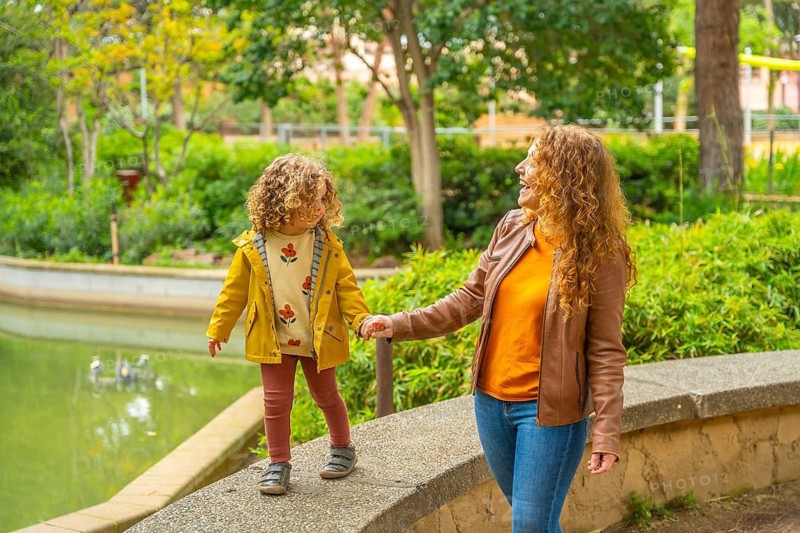 Girl walking on rail holding the hand of her mother along a park next to a pool