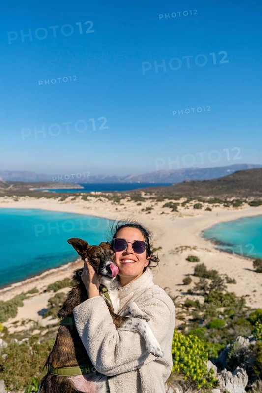 A woman in sunglasses and a cozy jacket holds her dog with love as they enjoy the sunny, picturesque beach landscape of Greece, surrounded by stunning turquoise waters