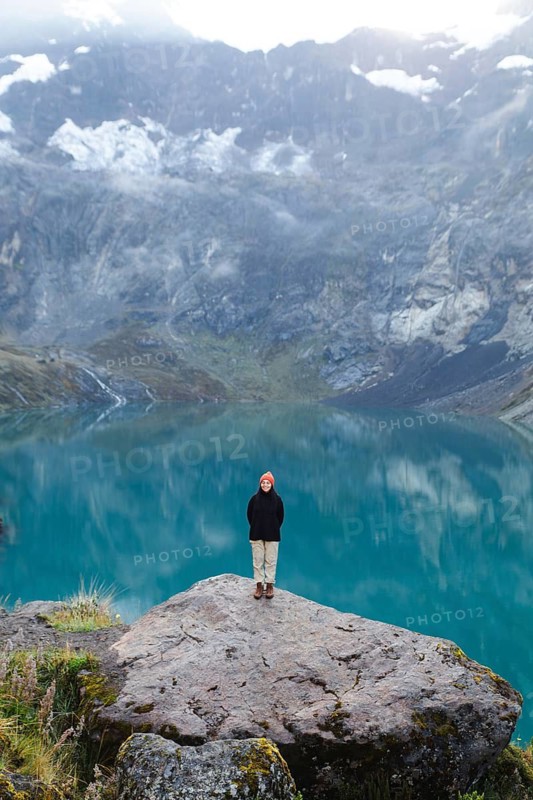 An Ecuadorian woman in a hat and jacket stands on a rock overlooking the turquoise lake at El Altar Volcano, Ecuador, with majestic mountains in the backdrop