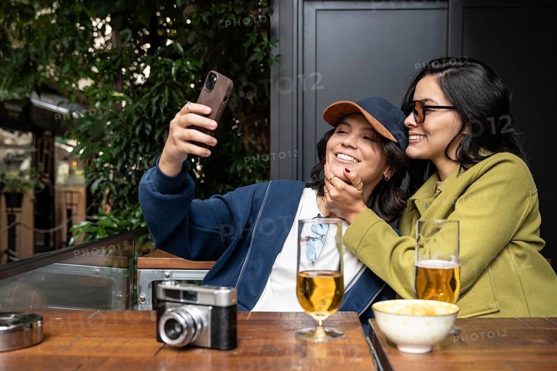 A joyful couple takes a selfie while enjoying drinks at an outdoor cafe. The relaxed atmosphere, filled with greenery, complements their laughter and affection