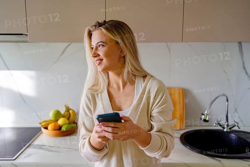 A woman with long blonde hair smiles while holding a phone in a modern kitchen. She wears a cozy sweater and stands near a sink with a bowl of fresh fruit