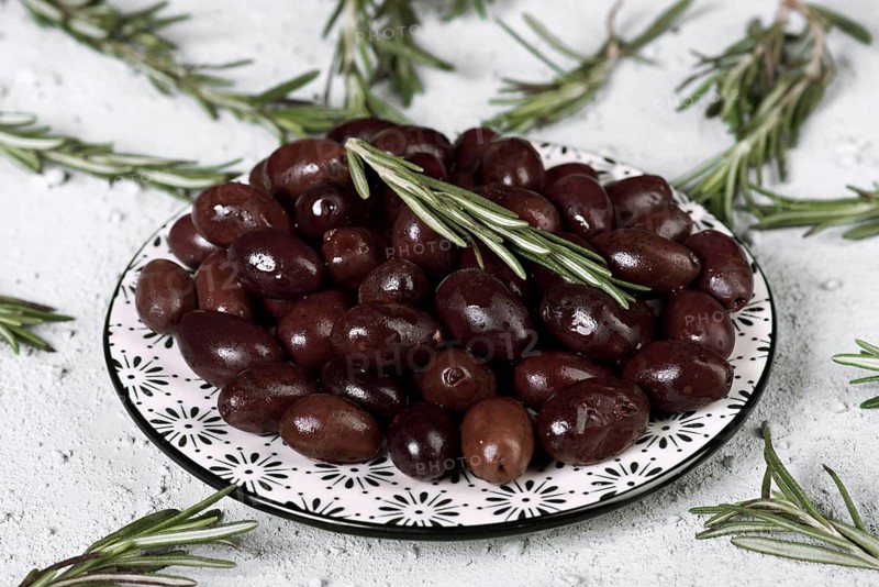 A decorative plate filled with black olives is garnished with sprigs of fresh rosemary. The serene arrangement rests on a textured surface, creating a rustic feel