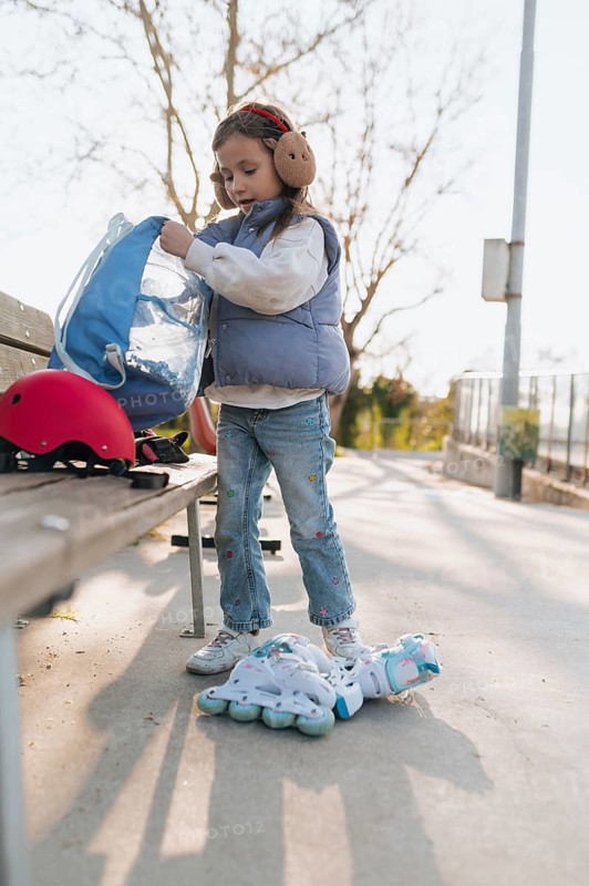 A young girl on roller skates is organizing her belongings in a blue bag at a park bench, ready for a skating adventure