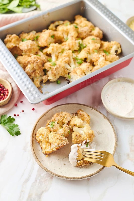 Top view of baked cauliflower in a metal baking tray. Each floret is coated in breadcrumbs and sprinkled with melted cheese and fresh herbs, accompanied by a creamy dip on a rustic ceramic plate