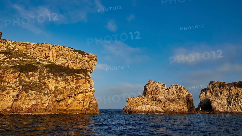 Cliffs with distinctive shapes rise out of the calm blue sea under a cloudy sky, morning light, Tsichli Babas islet, Navarino Bay, Pylos, Peloponnese, Greece