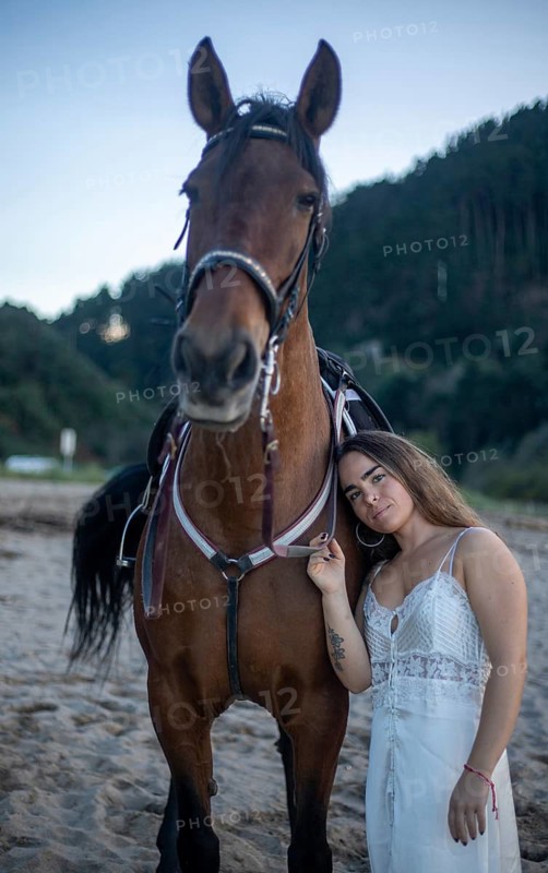 A young woman in a white dress stands beside her horse on a sandy beach. The duo is set against a backdrop of trees, capturing a serene moment at dusk