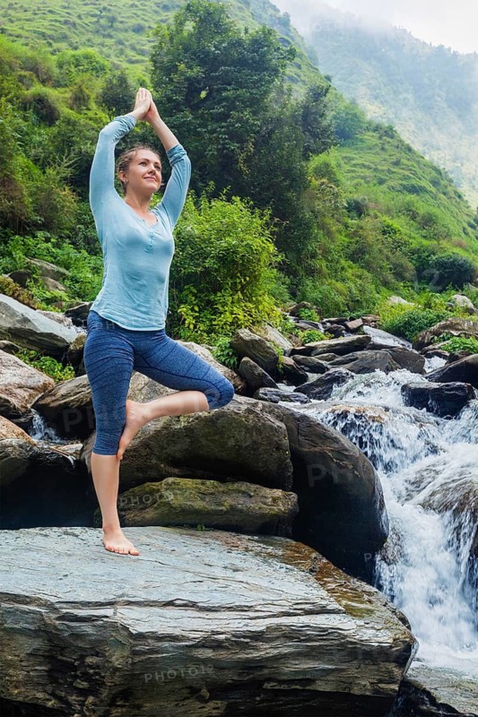 Woman in Hatha yoga balance yoga asana Vrikshasana tree pose at waterfall outdoors