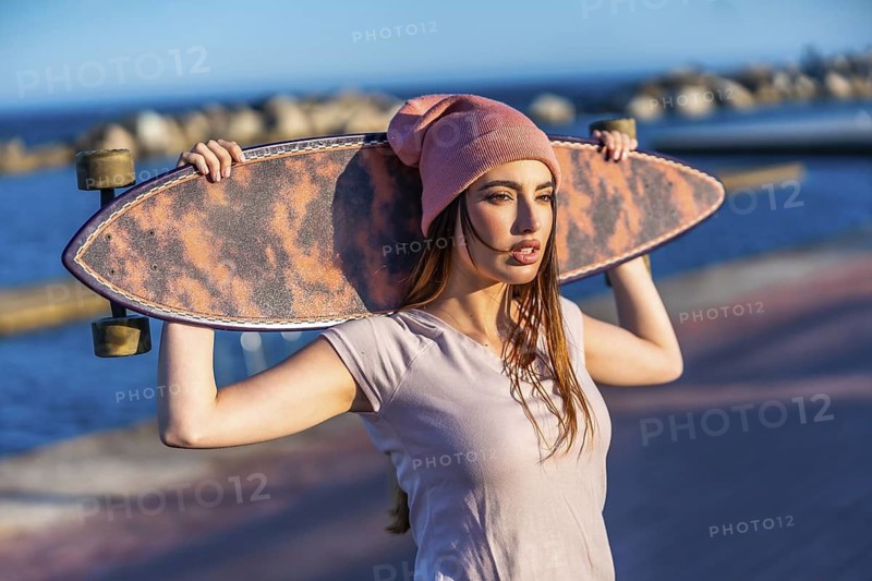 Young woman enjoying the sunset by the sea, holding a longboard on her shoulders, embodying freedom and a trendy, active lifestyle