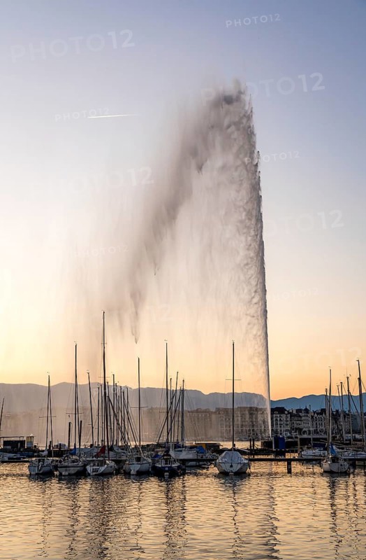 Iconic Jet d'Eau fountain in Geneva shoots water high into the air, backed by a sunset sky and silhouetted Alps. Moored sailboats add charm to the scenic view