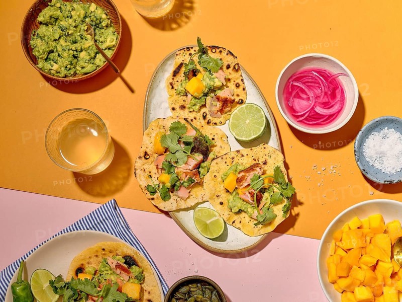 Top view of a taco meal preparation on a vibrant yellow and pink background. The image displays tacos filled with salmon, mango, coriander, and avocado. Additional sides include guacamole, pickled onions, lime wedges, and salt