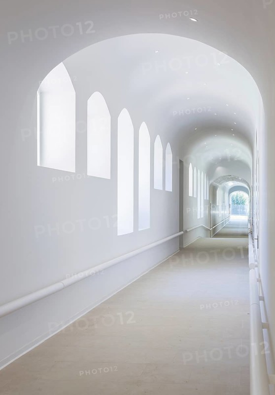A serene white corridor of hospital featuring high arched windows allowing abundant natural light to flood the space, creating a soft and peaceful ambiance as it stretches endlessly