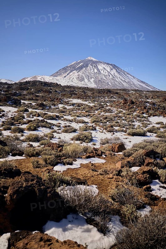 Teide volcanic mountain, partially covered in snow, rises under a clear blue sky. The rocky terrain in the foreground adds texture to this breathtaking landscape
