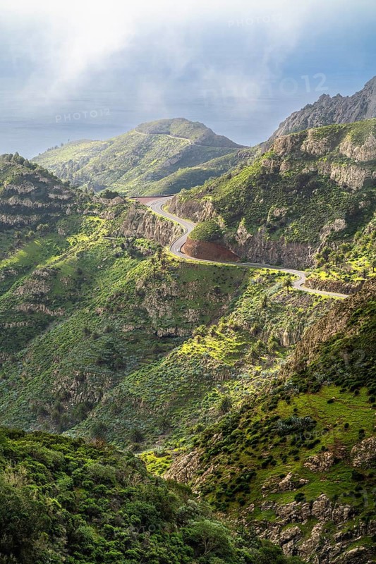 Landscape in the south-east of La Gomera near Vegaipala. Mountain slopes with palm trees and agaves, a road, sea, sun and clouds. La Gomera, Canary Islands, Spain