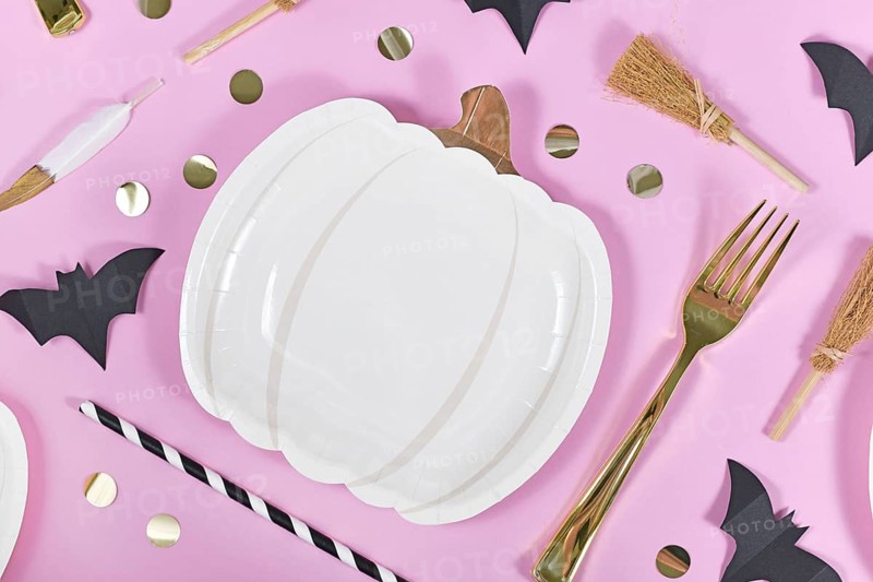 Halloween party flat lay with pumpkin shaped paper plates, bats, golden confetti, forks and spoons and witch brooms on pink background