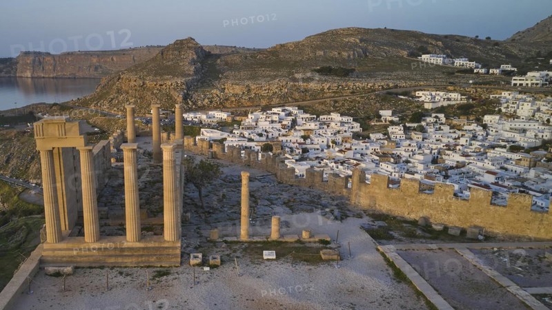 Drone shot, first morning light, Lindos, Acropolis of Lindos, Temple of Athena Lindia, Historical columns in the foreground with a coastal town of white buildings behind, Archaeological site, Lindos, Rhodes, Dodecanese, Greek Islands, Greece