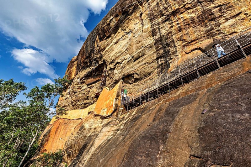 A narrow path leads along an imposing rock face over which a blue sky arches, The Lion Rock near Sigiriya in Sri Lanka